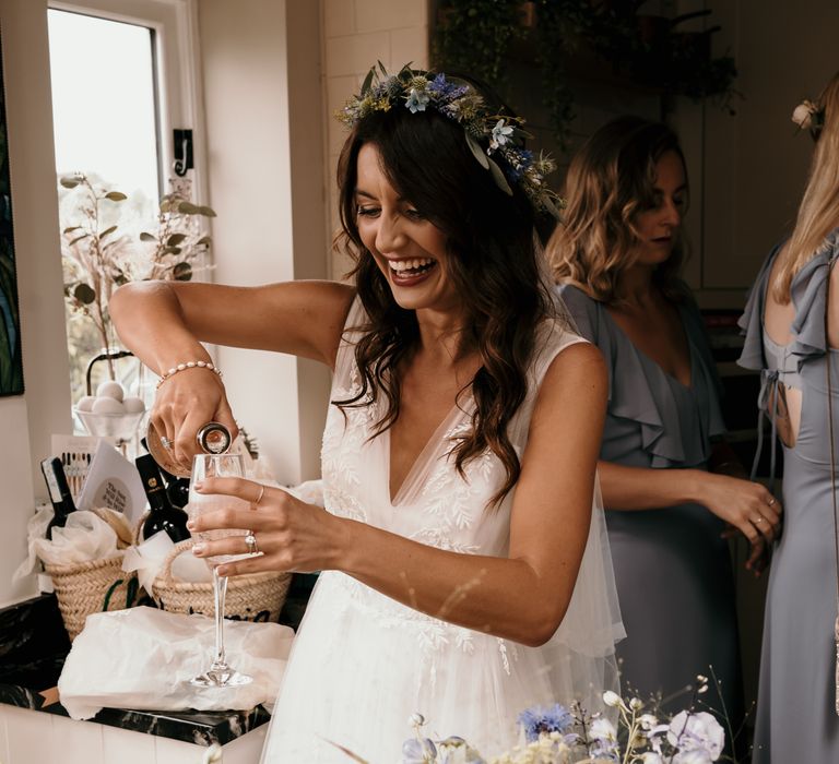 Dark haired bride pours champagne on the morning of her wedding whilst laughing and wearing floral crown