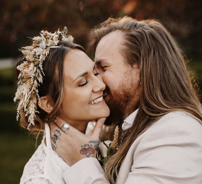 Groom in a beige suit with long hair and tattoo's kissing his bride's neck in a lace dress and dried flower crown