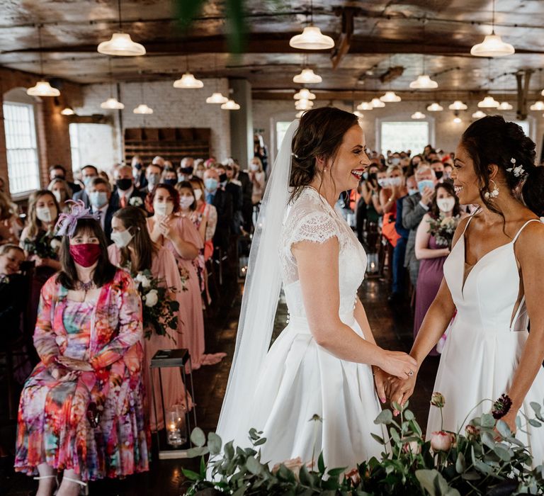 Smiling bride in white cami wedding dress with shoulder veils holds hands with smiling bride in lace capped sleeve wedding dress and veil in front of applauding guests during wedding ceremony at The West Mill Derby