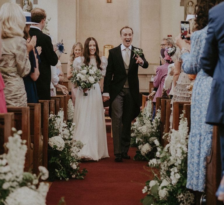 Bride in white lace puffed sleeved Daalarna wedding dress holding white and green bridal bouquet walks down the church aisle holding hands with groom after being married at church in Surrey