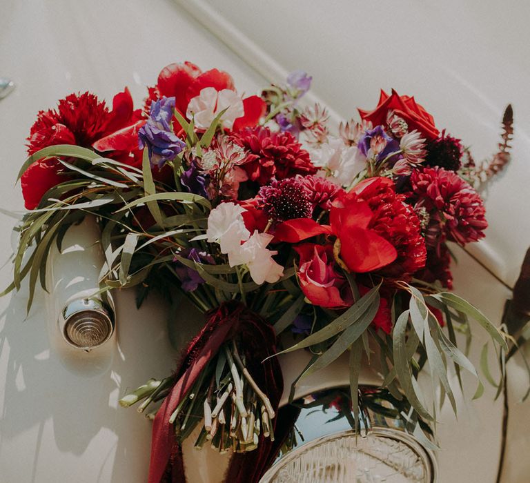 Deep red and purple jewel tone wedding bouquet sitting on a vintage white wedding car