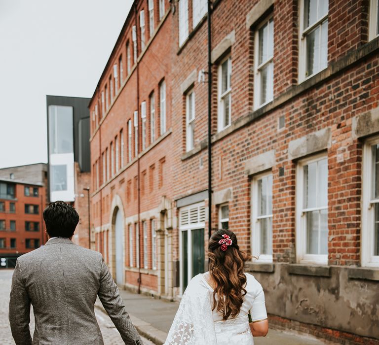 A bride a groom walk away from camera holding hands. He wears a check grey suit and she wears a white sari.