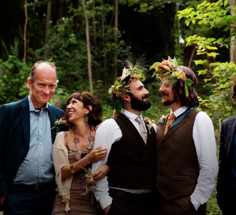 Grooms look lovingly at one another as they stand with their family outdoors within the woodland