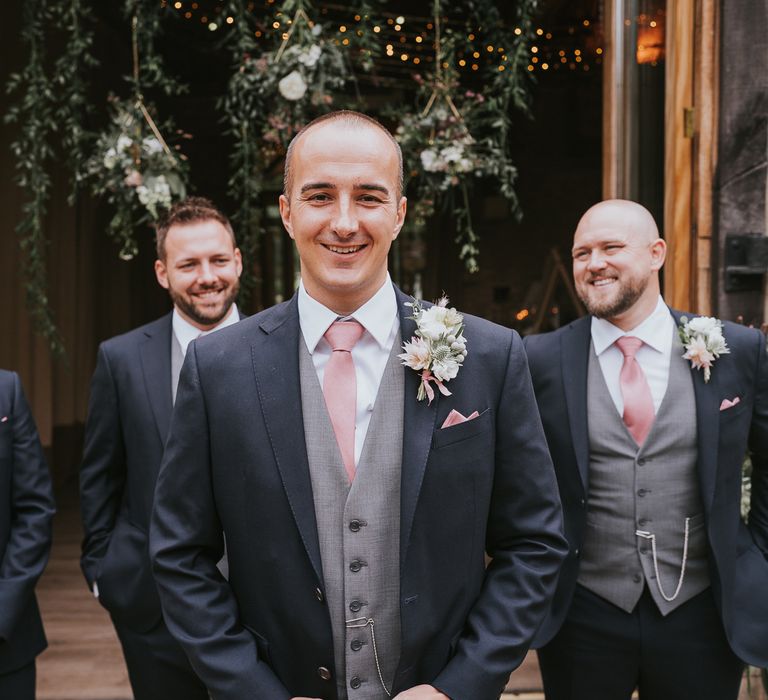 Groom and groomsmen in dark three piece suits with grey waistcoats, pink ties and protea and thistle buttonholes at Tythe Barn wedding with barn wedding flowers