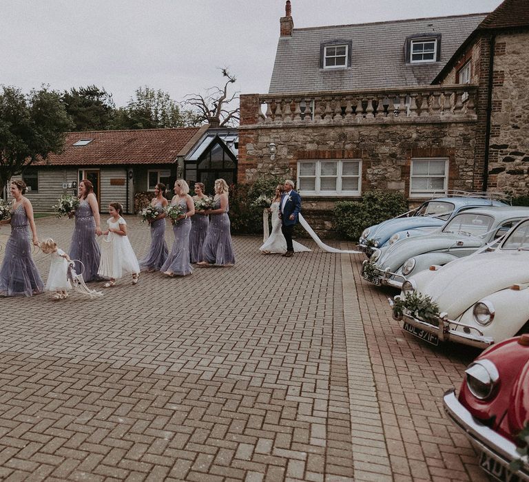 Bridesmaids in blue beaded cami bridesmaids dresses and flower girls in white dresses walk across courtyard followed by bride in Grace Loves Lace wedding dress and veil arm in arm with father in blue suit at Isle of Wight wedding