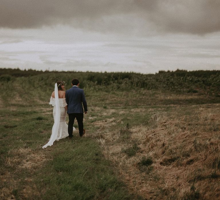 Bride in Grace Loves Lace bardot wedding dress, veil and flower crown walks arm in arm with groom in blue suit jacket walk through field at Isle of Wight wedding with macrame wedding decor