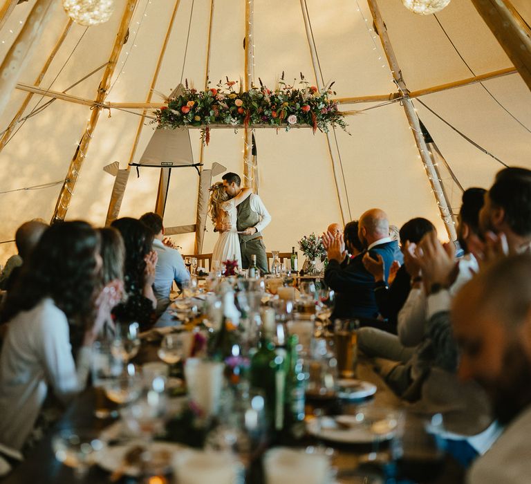 Bride and groom hug as guests sat at long wooden table applaud during tipi wedding breakfast at Wellington Wood Norfolk