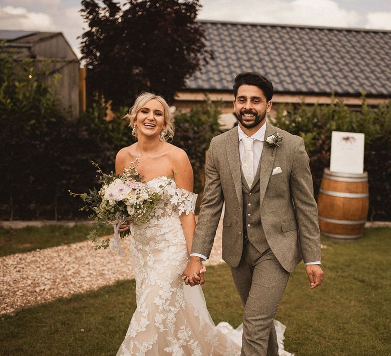 Bride in off the shoulder Enzoani wedding dress holding white, pink and green bridal bouquet walks holding hands with groom in grey three piece suit with white ties and floral buttonhole at Inkersall Grange Farm wedding