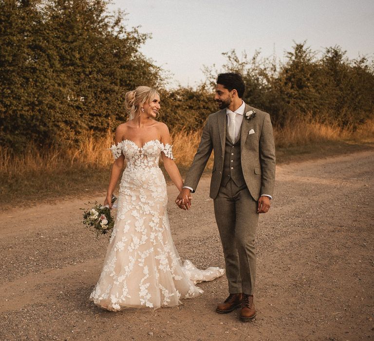 Bride in off the shoulder Enzoani wedding dress with train holds white and pink bridal bouquet as she walks down country lane holding hands with groom in grey three piece suit with white buttonhole at Inkersall Grange Farm wedding