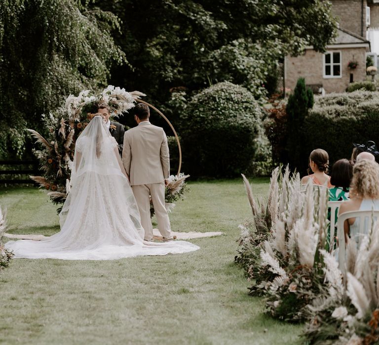 Bride in homemade wedding dress and veil stands at floral Moorgate altar with groom in beige suit during DIY garden wedding ceremony in Bedfordshire