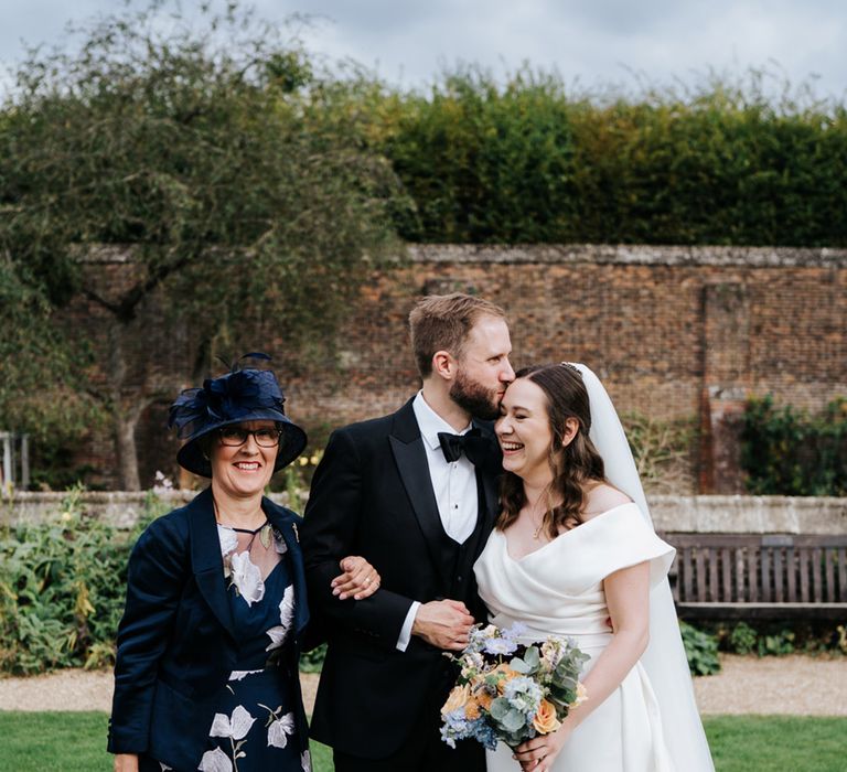 Groom kisses the top of his brides head as they stand outdoors for family pictures