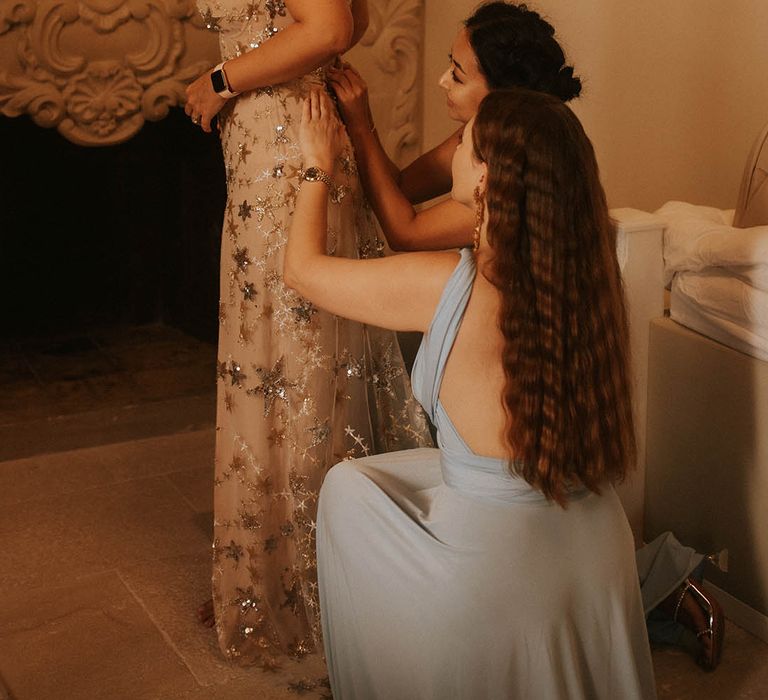 Bridesmaids kneel behind bride whilst doing up her wedding gown