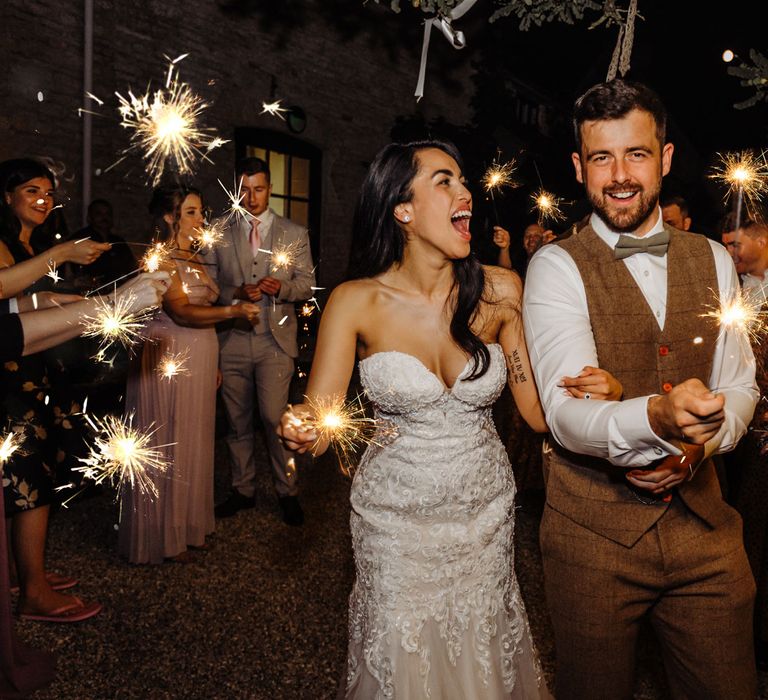 Bride in lace and tulle wedding dress with sweetheart neckline walks with groom in brown tweed waistcoat as they have sparkler exit