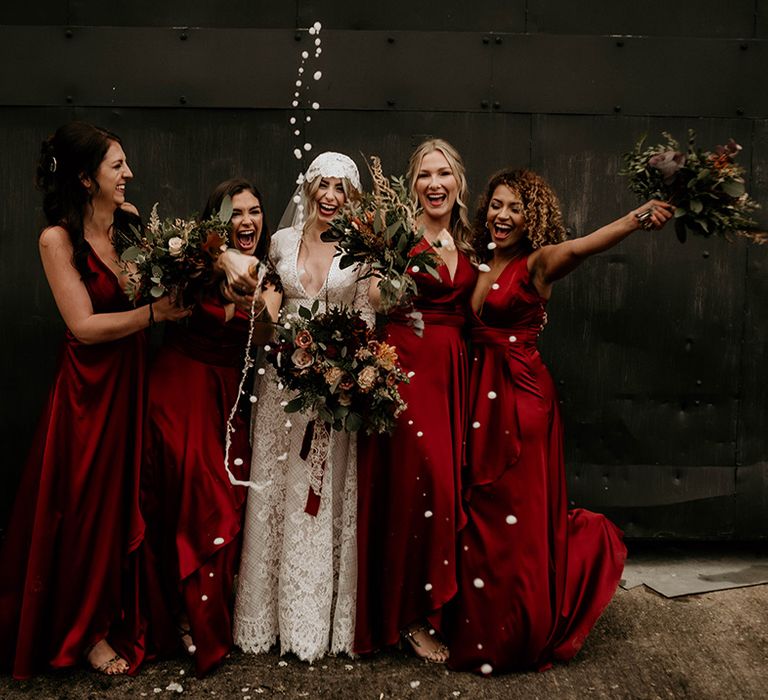 Fun bridal party portrait with bride in a lace wedding dress and bridal party in satin red bridesmaid dresses spraying champagne 