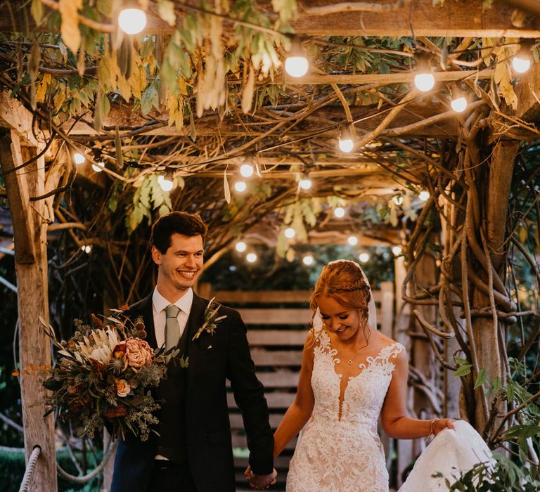 Bride in deep v lace wedding dress holding tulle train walks under Edison bulb canopy with groom in sage green tie holding mixed bridal bouquet