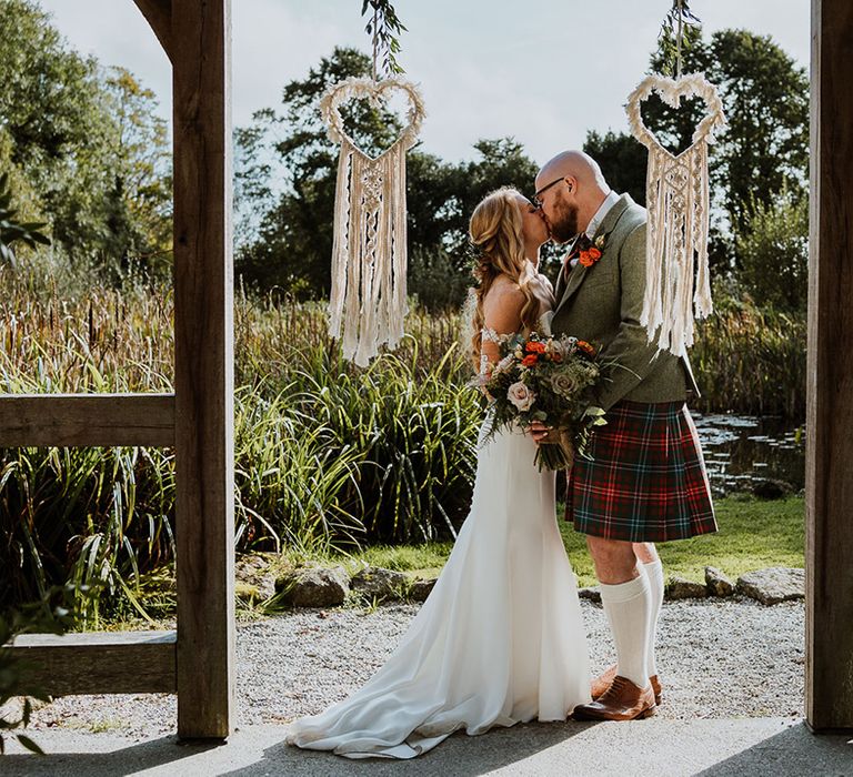 Bride & groom kiss underneath macrame bunting outdoors 