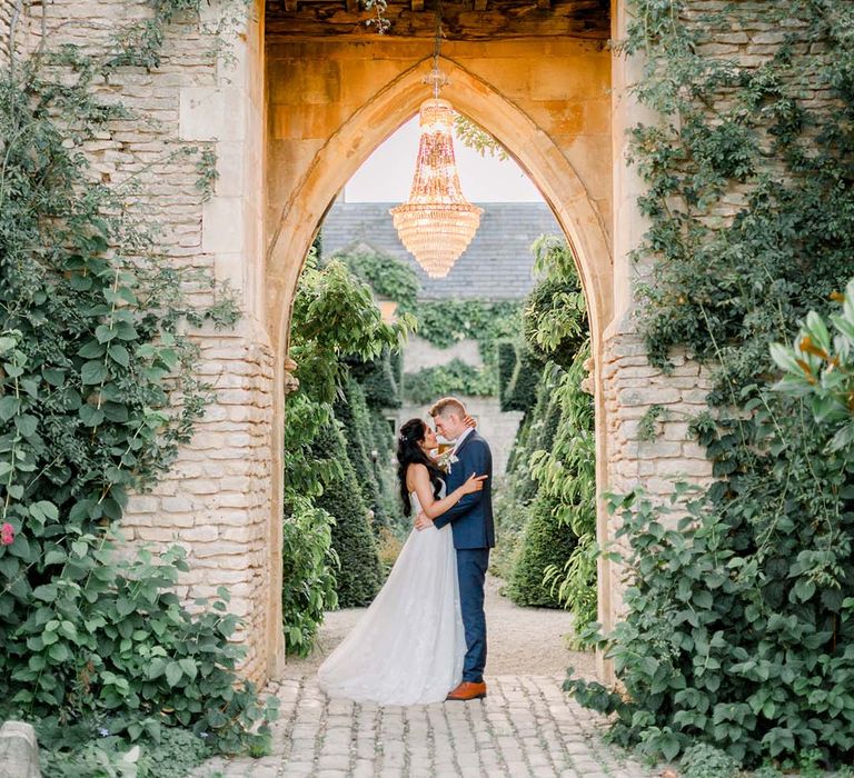 Bride & groom kiss under archway outdoors with chandelier hanging above them on their wedding day