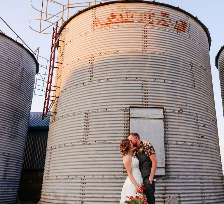 Bride in lace wedding dress and roses in her hair kisses groom in short sleeve patterned short by grain store for festival wedding