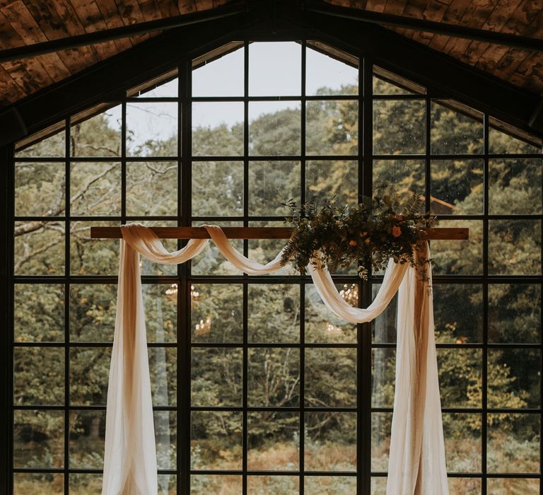 Wooden wedding arch with white draping, floral cloud and candles in front of large window at the Hidden River Barn wedding venue