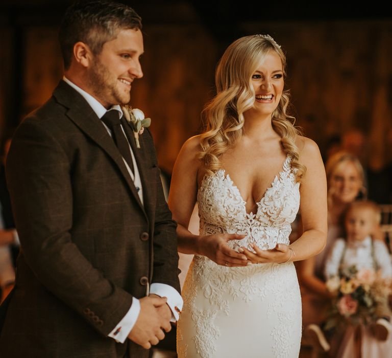 Smiling bride with curled hair in lace wedding dress stands with groom in brown three piece suit and buttonhole during rustic barn wedding ceremony