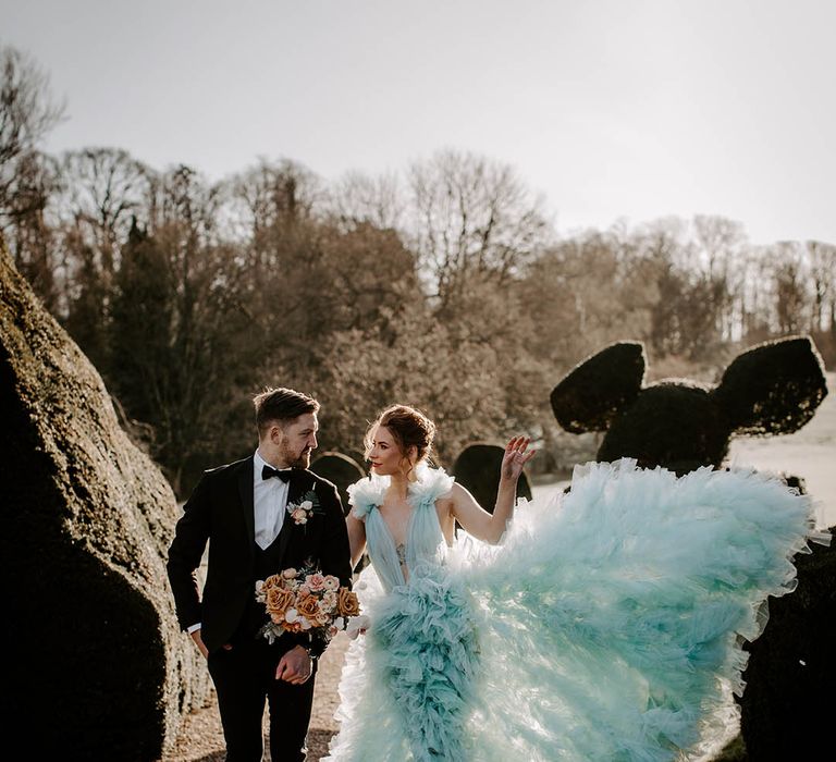 Bride in a ruffle light blue wedding dress fanning out the skirt as she walks arm in arm with her groom in a tuxedo at Newburgh Priory