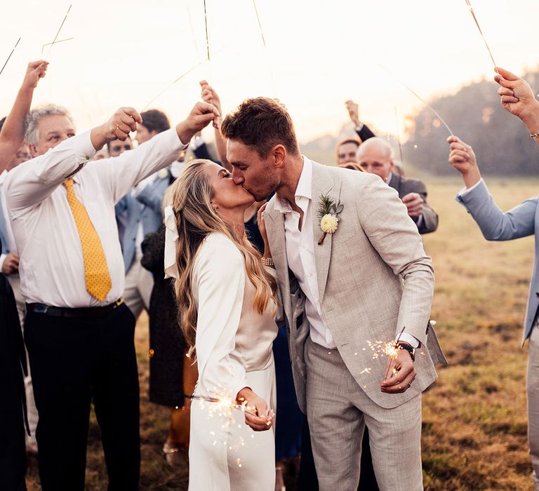 Bride in satin dress with train and long sleeve wrap top kisses groom in grey suit as they hold sparklers