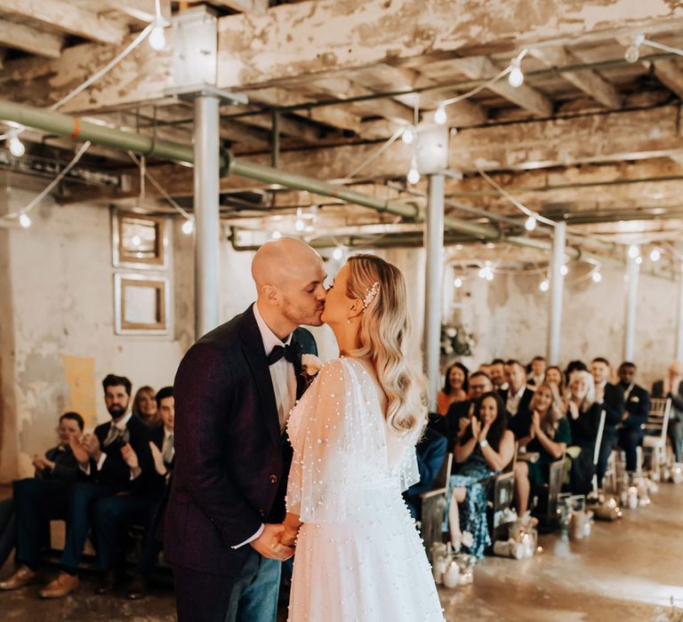 Groom in navy tweed suit with brown shoes kisses bride in pearl wedding dress with train during wedding ceremony at Holmes Mill