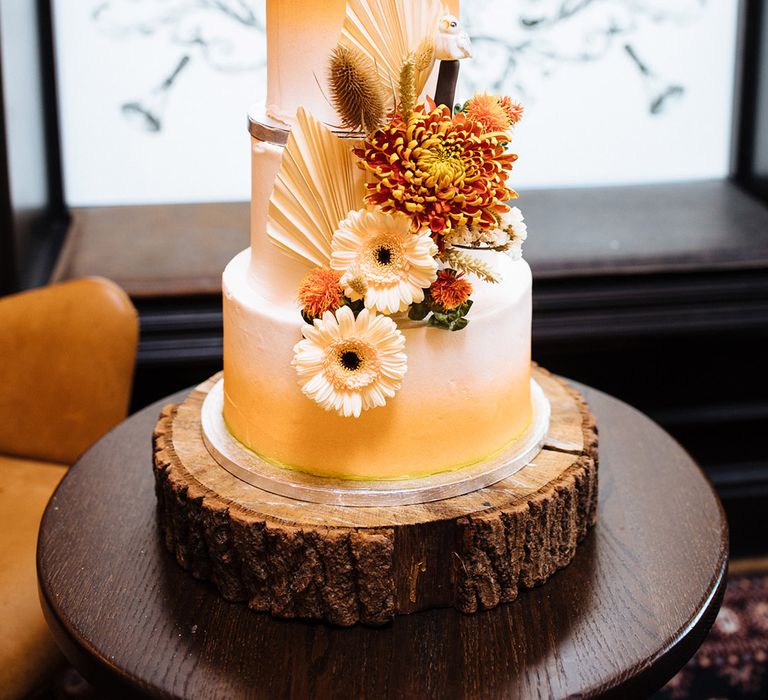 Three-tier wedding cake with orange ombré frosting with orange and beige flowers on tree trunk stand and custom cake toppers 