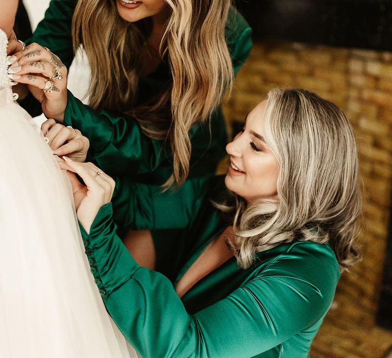 Bridesmaids in emerald green long sleeve dresses and French tip nails do up buttons on the bride's wedding dress