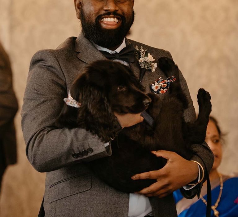 Groom in dark grey suit and white neutral boutonniere holding black puppy 