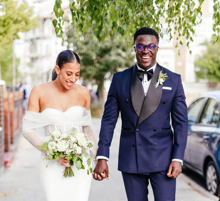 Smiling groom in black tie walks holding hands with bride in off the shoulder corset style fitted wedding dress with white flower bouquet