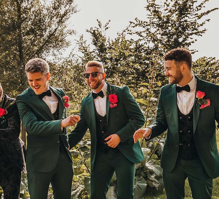 Groom with groomsmen in emerald green and black patterned suits with red rose boutonnières as they all smile