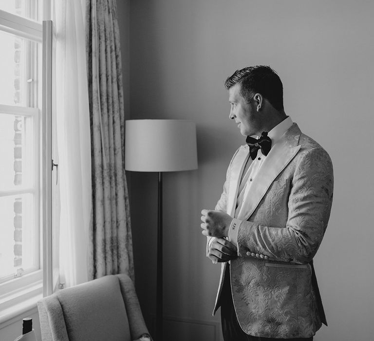 Groom fixes his cufflinks as he looks out the window in custom tuxedo black tie for his destination wedding in London 