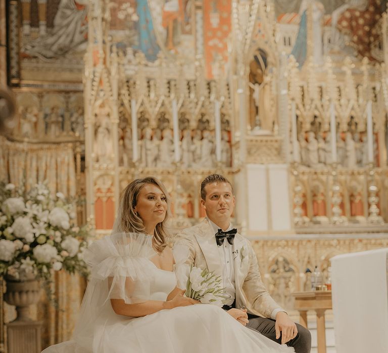 Bride in Halfpenny London wedding dress sits with groom at the altar holding bouquet of white tulips