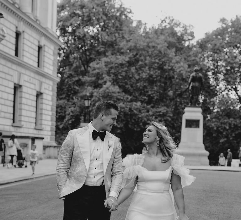 Bride in ruffle sleeves and skirt wedding dress holds white tulip wedding bouquet and hands with the groom in black tie 