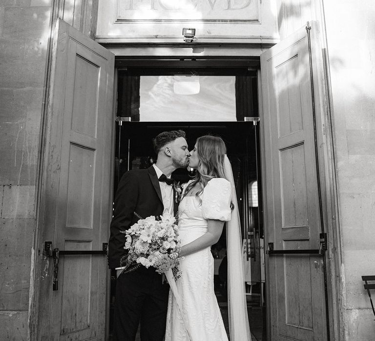 Bride and groom share a kiss outside the main doors of their wedding reception venue 