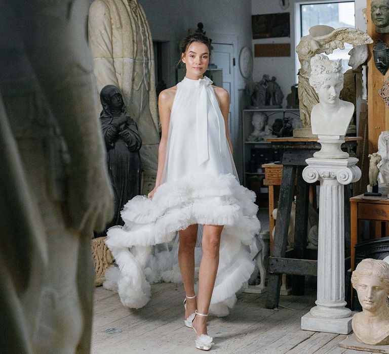 Bride in high neck wedding dress with ruffle detail and a bow wearing pointed white wedding shows with white flowers