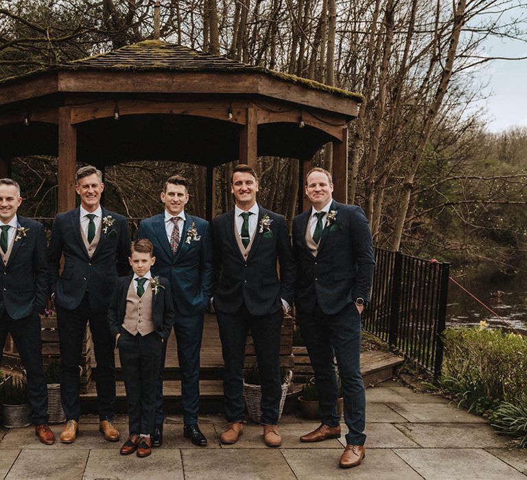 Groomsmen in blue suits with beige waistcoats and green ties stand with groom in blue suit and floral tie