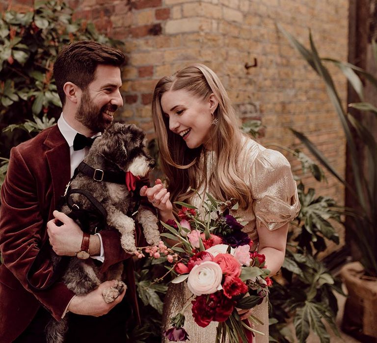 Groom in burgundy suit jacket with bride in gold wedding dress holding their pet dog wearing red bow tie collar 