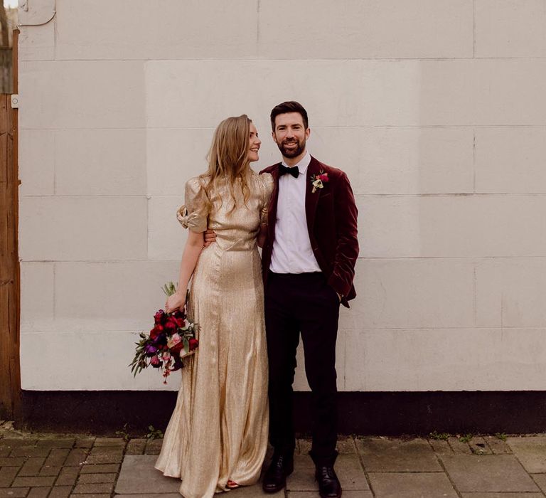 Groom wearing black tie and red velvet suit jacket has his arm around the bride in sparkly gold wedding dress holding dark wedding bouquet