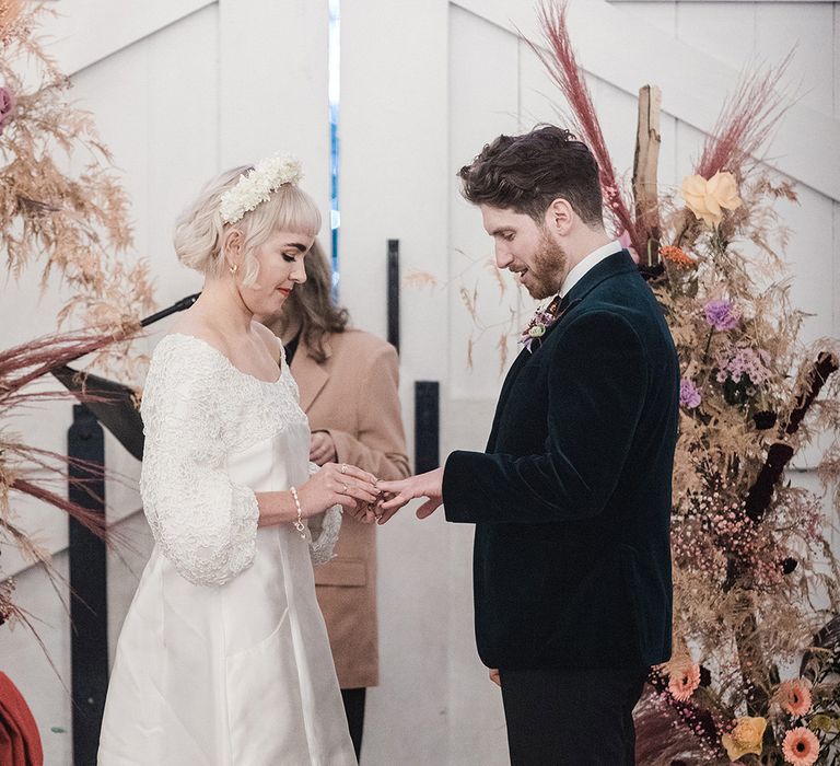 Bride in short wedding dress with white flower headband places the ring on the groom's finger at wedding ceremony