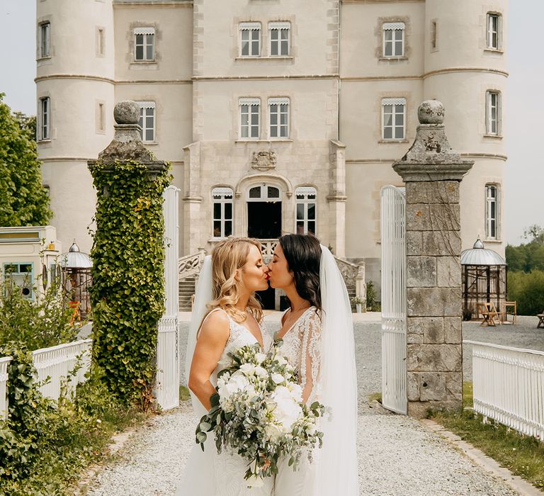 Brides share a kiss both wearing red lipstick in front of the chateau 