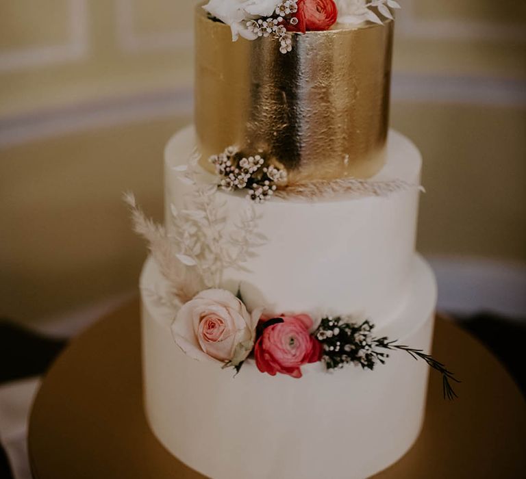 Four tier white frosted wedding cake with one layer frosted in gold decorated with pink and white flowers and dried grass