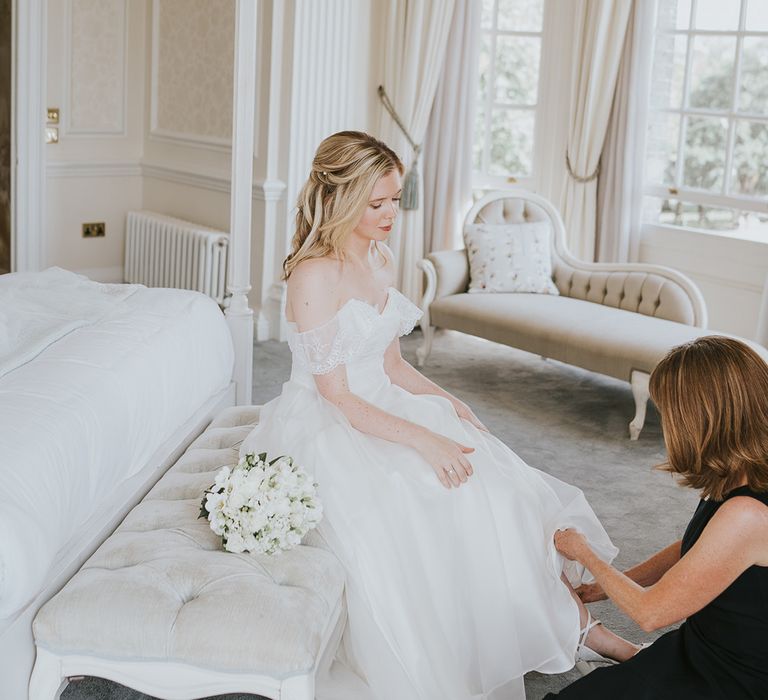 Bride in off the shoulder dress has white flower wedding bouquet gets help putting her shoes on for her wedding day 