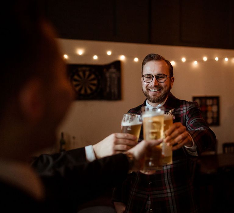 Groom holds beer up to friends in cheers as he wears bespoke tartan suit on the morning of his wedding day