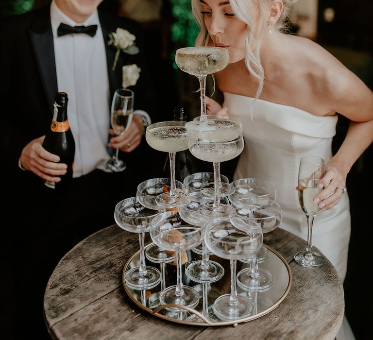 Bride sips from the champagne tower with groom in black tie watching