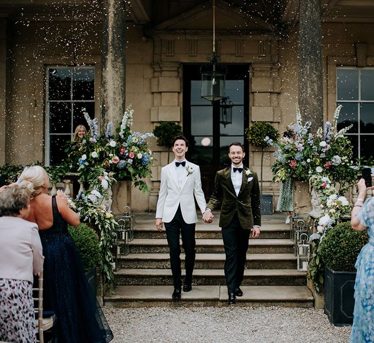 Groom in white black tie holds the hand of his groom wearing green velvet jacket as they walk outside Birdsall House as confetti falls around them