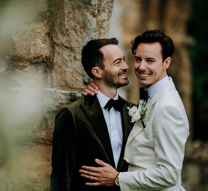 Groom wearing white jacket leans in to his groom wearing velvet green jacket and black tie 