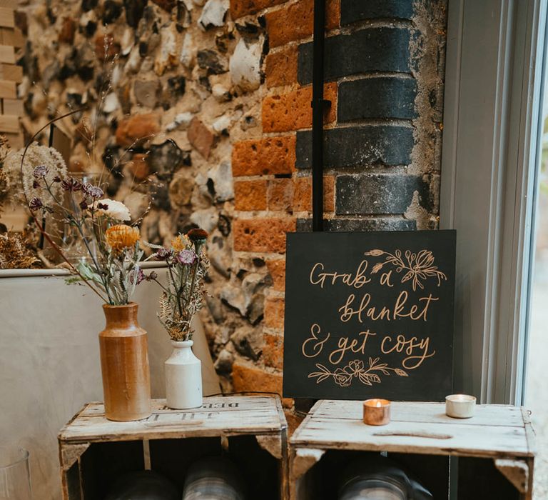 Wooden crates filled with blankets for wedding guests with tea lights, a black sign and autumnal flowers 