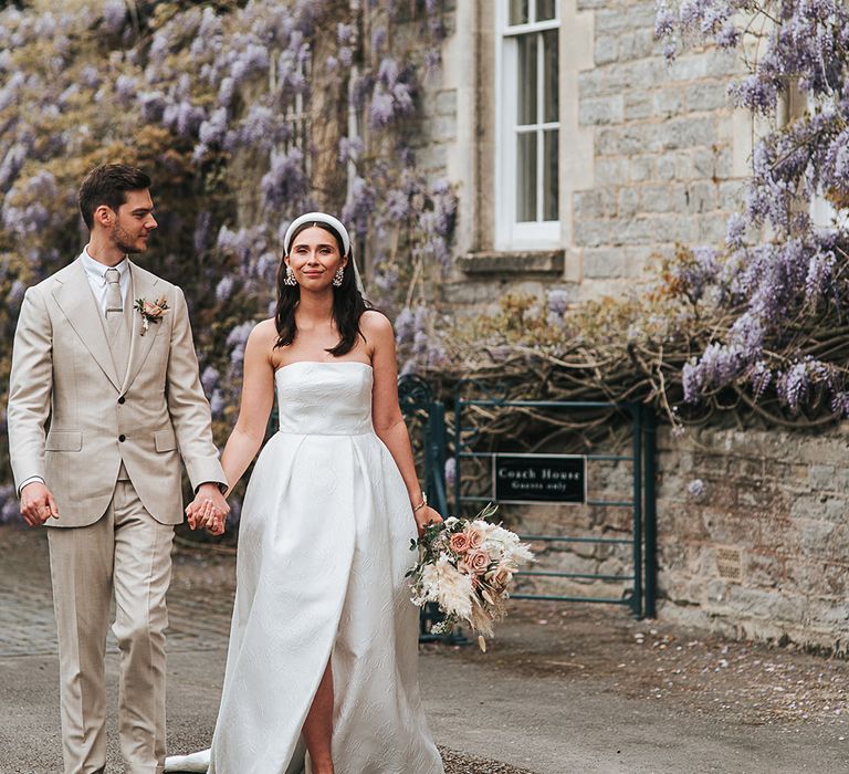 Groom in a neutral wedding suit walks hand in hand with the bride in a strapless brocade pleated wedding dress and headband 